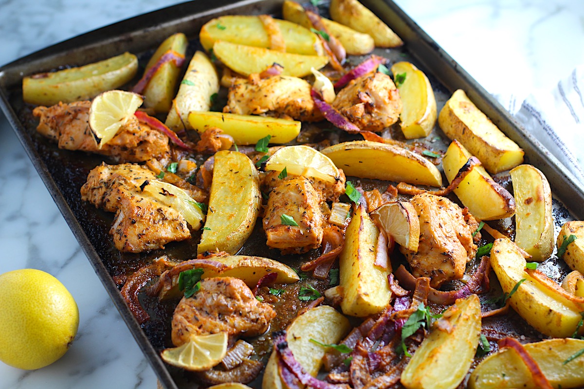 Crispy, golden brown Greek Lemon Chicken and Potatoes with red onions and fresh parsley garnish on a sheet pan on the counter. Whole lemon sitting to the left on the counter.