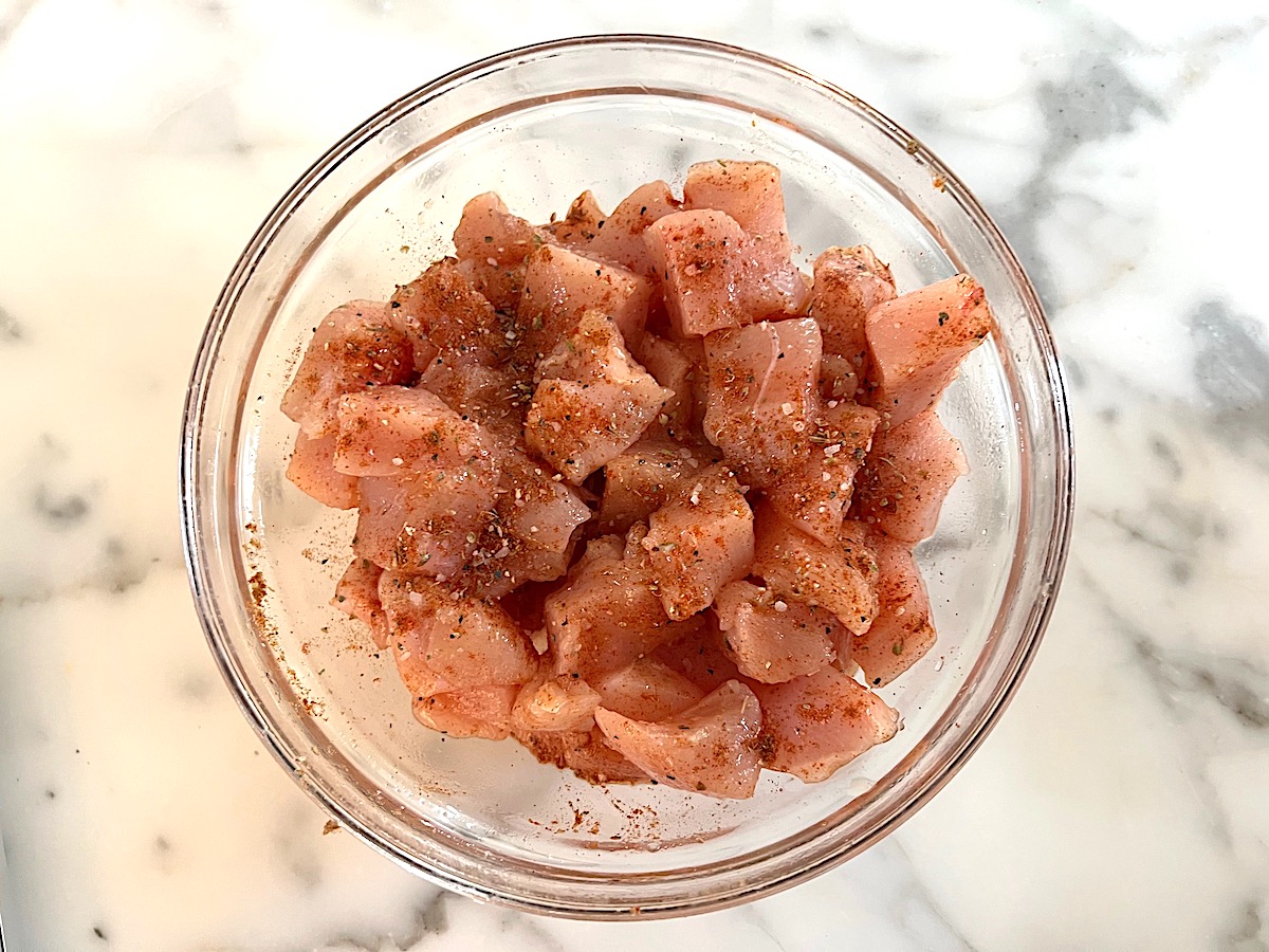 Cubed raw chicken breast in a clear bowl seasoned for Semi-frozen chicken sliced on a cutting board for Chicken Chorizo Orzo.