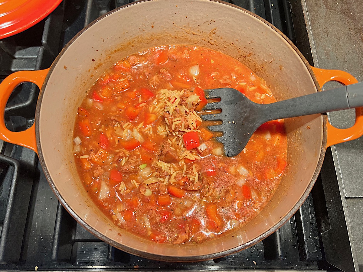 Broth, tomato paste, and orzo added and cooking in a pot on stove for Chicken Chorizo Orzo.
