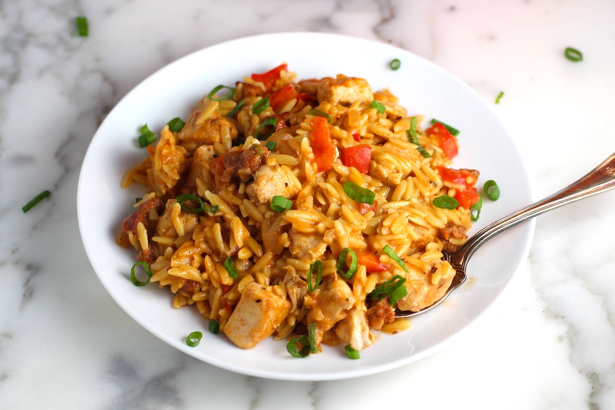 Creamy Chicken Chorizo Orzo dinner with red peppers served on a plate with sliced scallions sprinkled over top and on the plate on the counter. A fork is resting on the plate with a scoop of the orzo and chicken.