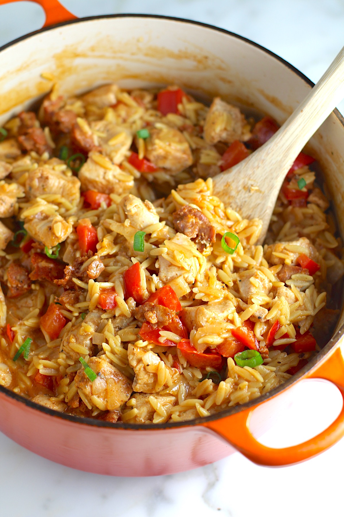 Creamy Chicken Chorizo Orzo dinner with red peppers in a dutch oven sitting on the counter with a wooden spoon scooping.