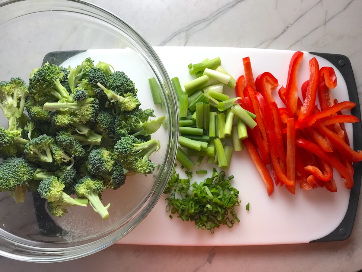 Broccoli florets in a bowl next to scallions and red peppers cut into pieces on a cutting board for Beef and Broccoli Noodlle recipe.
