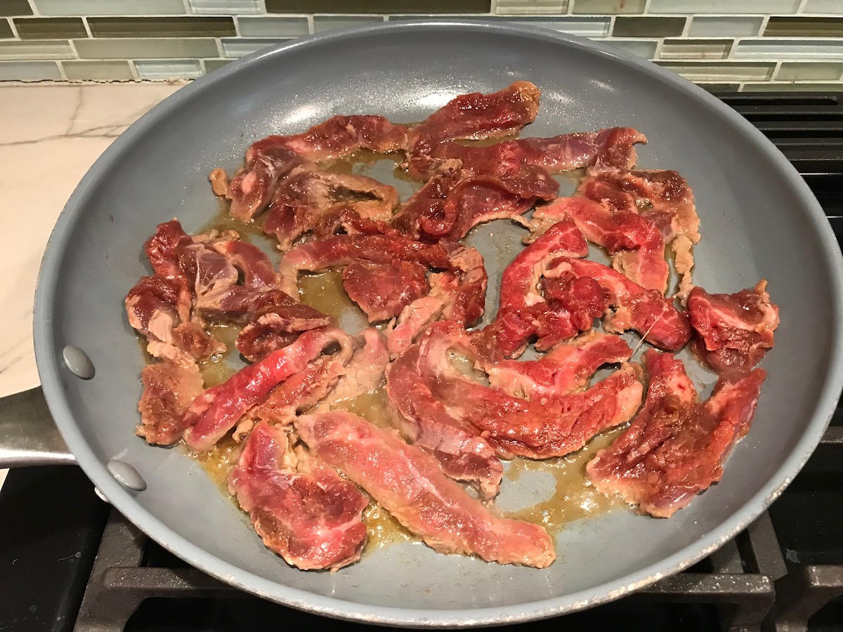 Raw flank steak strips being seared in a frying pan for beef and broccoli noodles recipe.