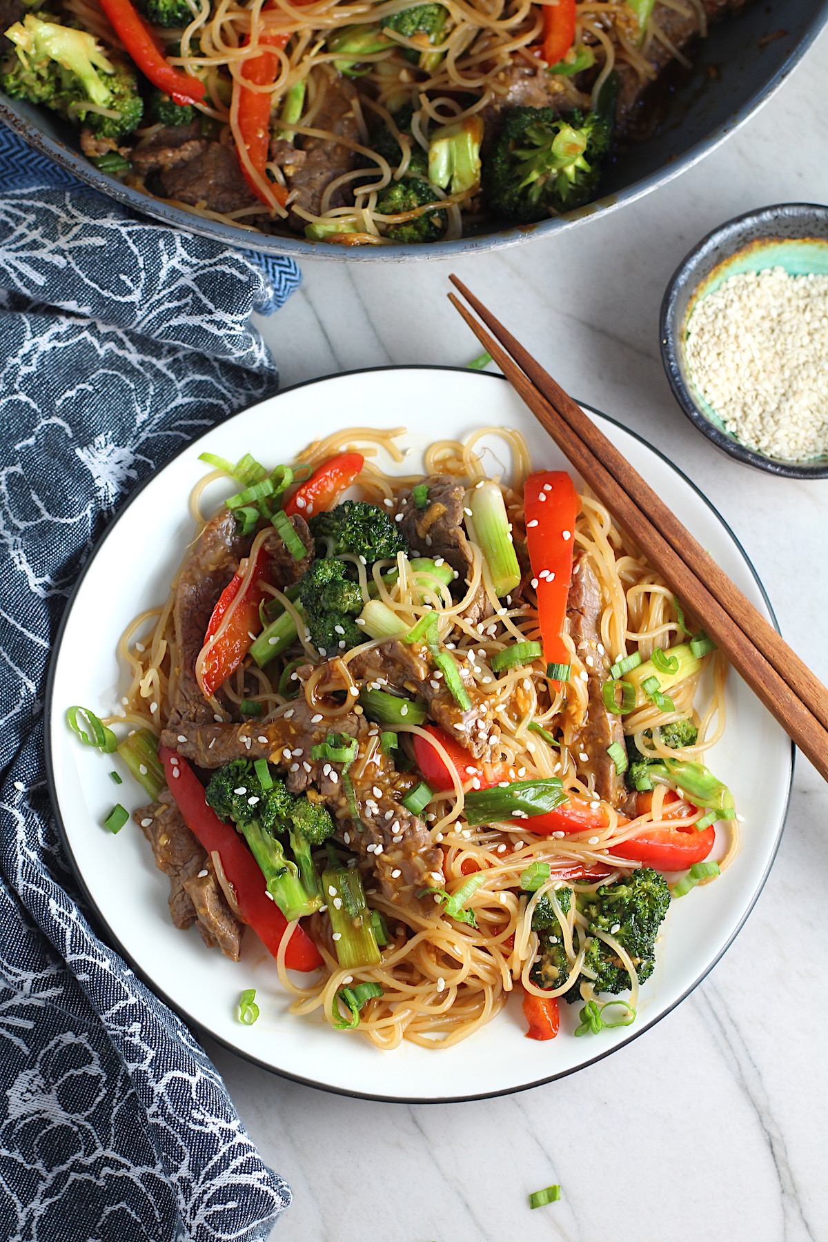 Steak, broccoli, red pepper, scallions, and noodles stir fried and on a plate with sesame seeds on top and chopsticks resting on the side for Beef and Broccoli Noodles recipe.