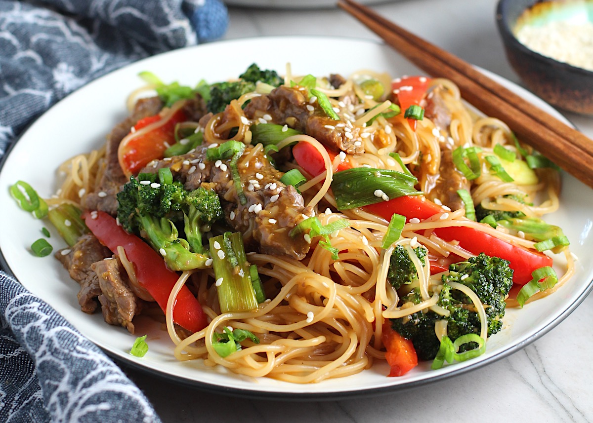 Steak, broccoli, red pepper, scallions, and noodles stir fried and on a plate with sesame seeds on top and chopsticks resting on the side for Beef and Broccoli Noodles recipe.