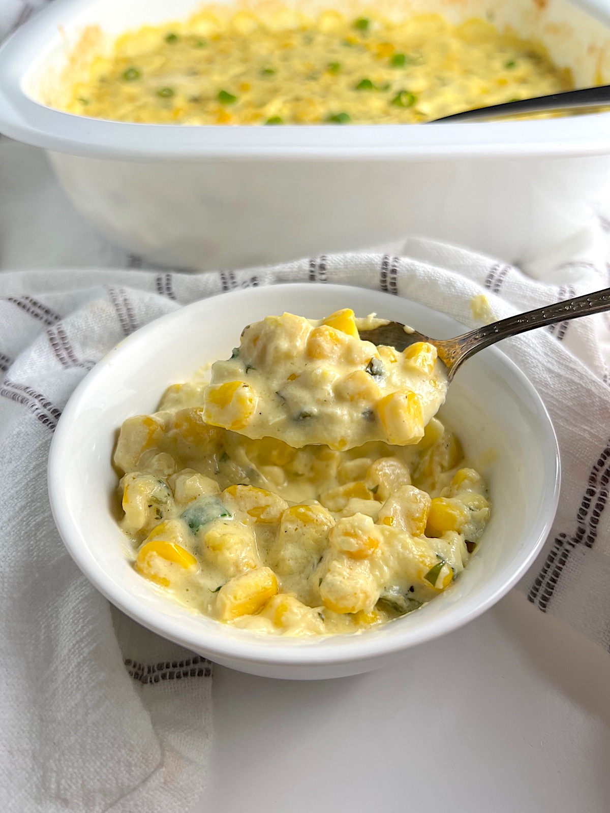 Creamy Mexican Street Corn Casserole in a bowl with spoon scooping a bite on blue and white striped towel with casserole dish in background.