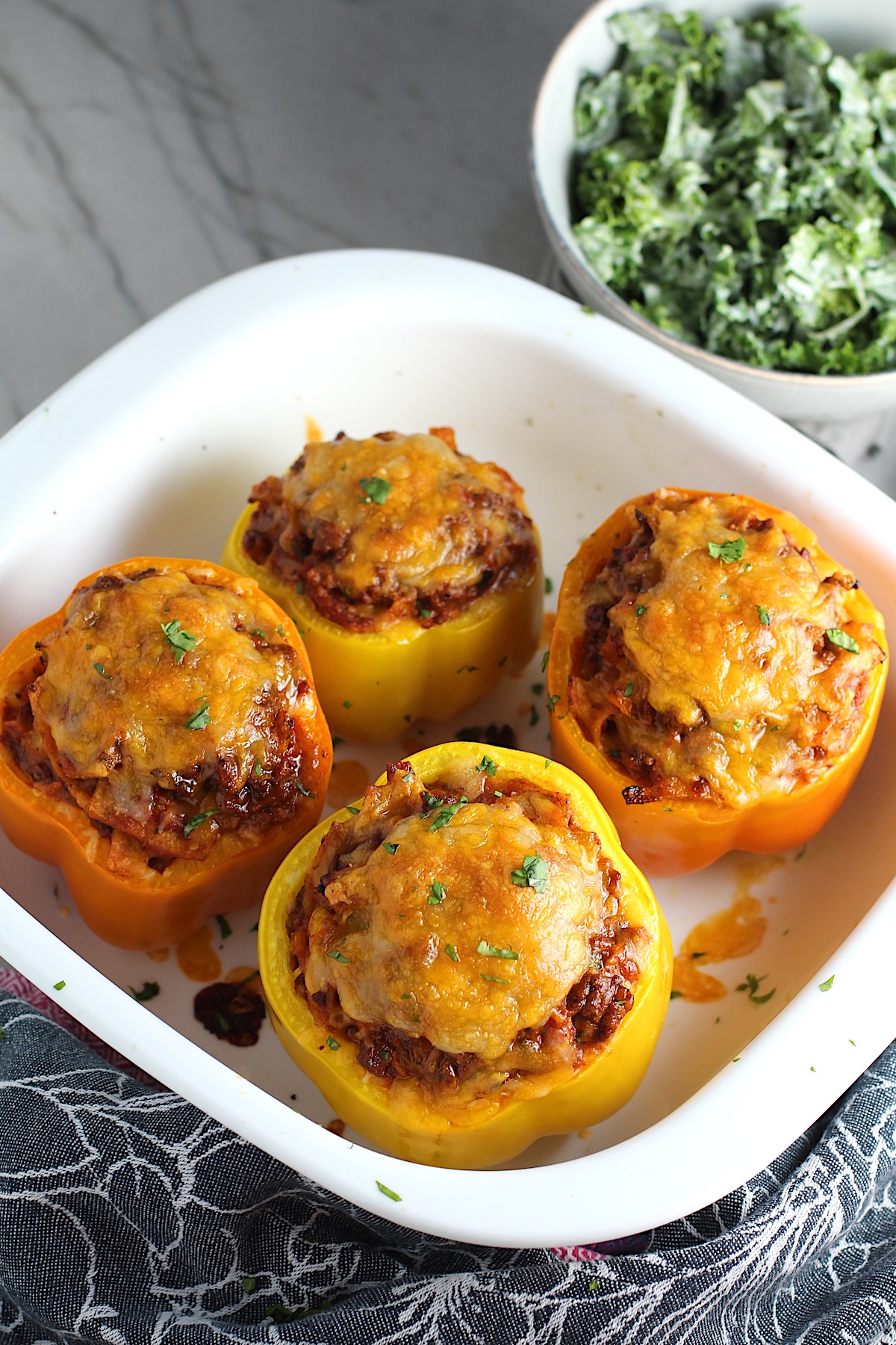 Four Tex Mex Beef Stuffed Peppers without Rice, but covered in melted cheese in a white casserole dish. Everything is set on the counter with a towel in front and a kale salad dressed in a creamy dressing in a bowl in the background.