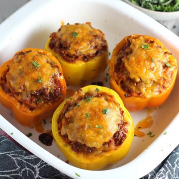 Four Tex Mex Beef Stuffed Peppers without Rice, but covered in melted cheese in a white casserole dish. Everything is set on the counter with a towel in front and a kale salad dressed in a creamy dressing in a bowl in the background.