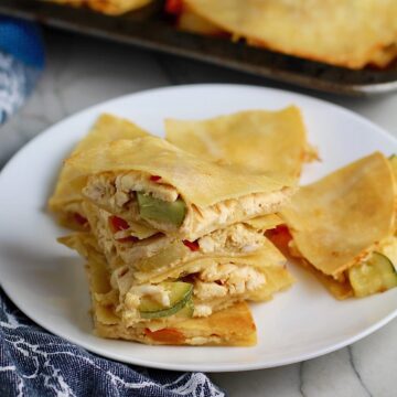 Stacked Veggie, Cheese, and Rotisserie Chicken Quesadillas on a plate on the counter next to cloth napkin with sheet pan holding quesadillas in the background.