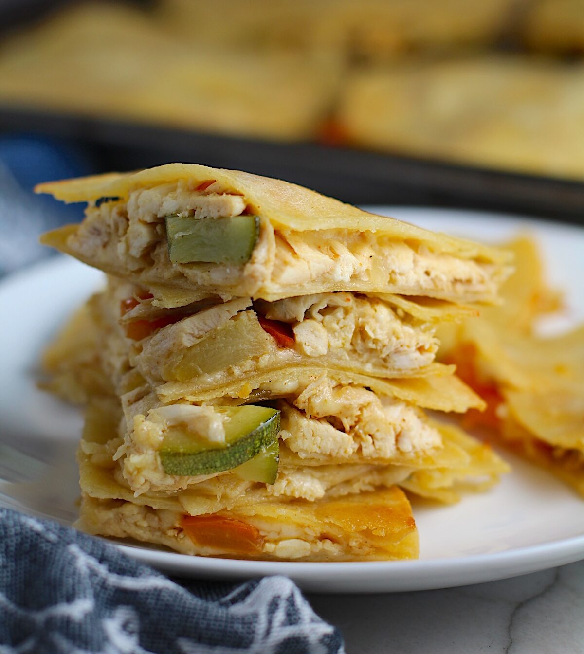 Stacked Veggie, Cheese, and Rotisserie Chicken Quesadillas on a plate on the counter next to cloth napkin with sheet pan holding quesadillas in the background.