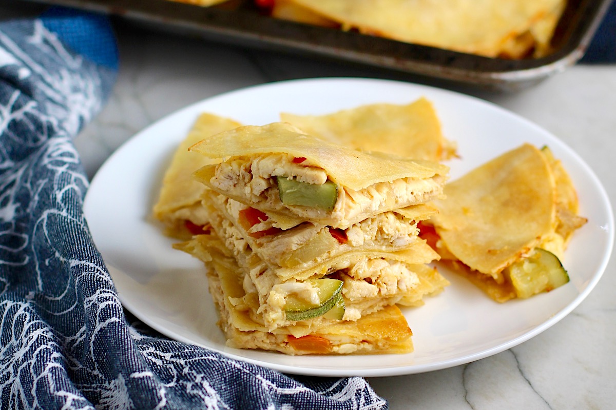 Stacked Veggie, Cheese, and Rotisserie Chicken Quesadillas on a plate on the counter next to cloth napkin with sheet pan holding quesadillas in the background.