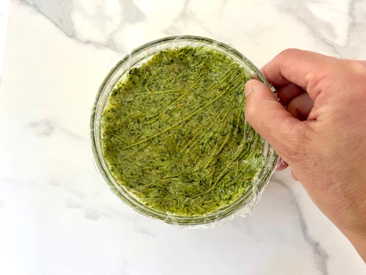 Hand sealing plastic wrap over finished pesto in clear bowl for Pesto Pasta with Tomatoes.
