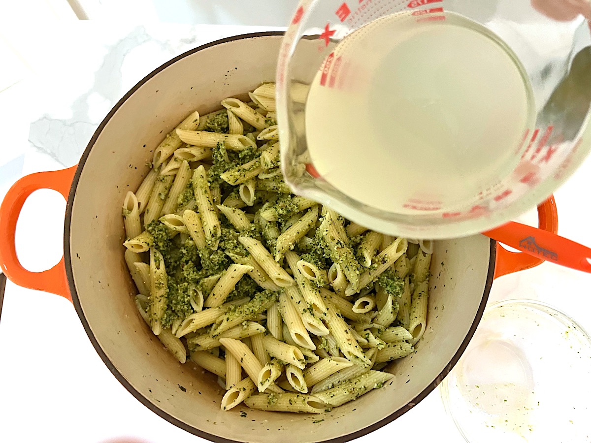 Measuring cup with pasta water over being poured over pasta mixed with pesto in pot for Pesto Pasta with Tomatoes.