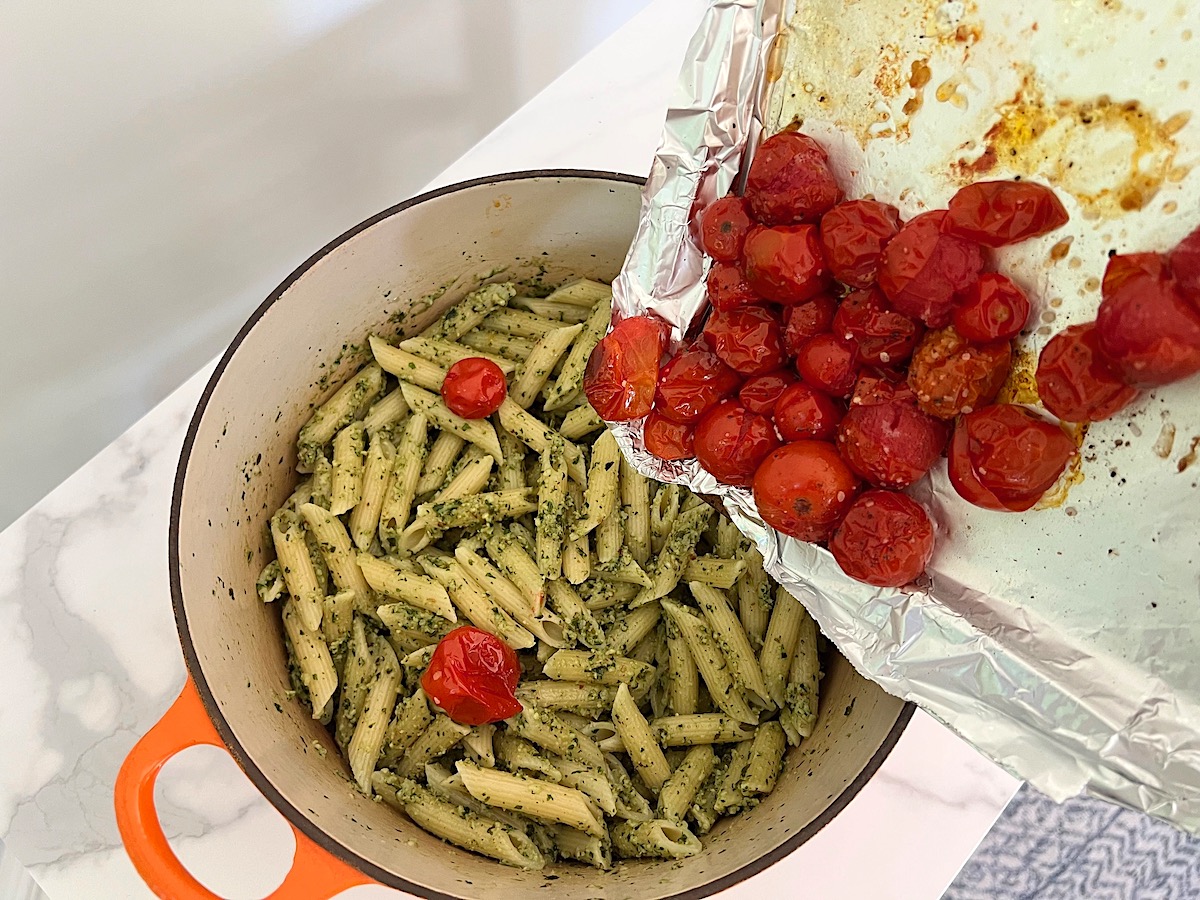 Roasted cherry tomatoes being added to pasta mixed with pesto in pot for Pesto Pasta with Tomatoes.
