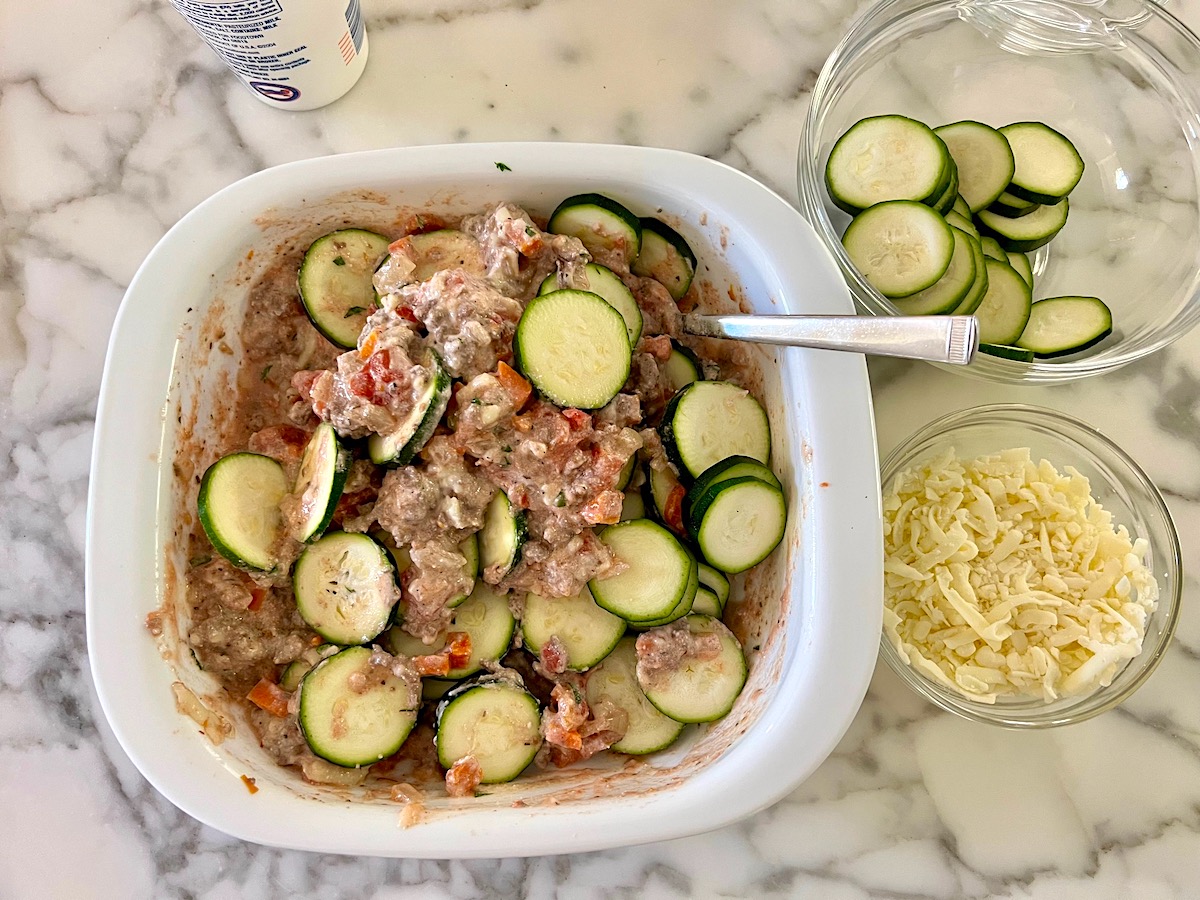 Raw zucchini slices being mixed into beef, tomato, and cheese mixture in casserole dish for Ground Beef and Zucchini Casserole recipe.