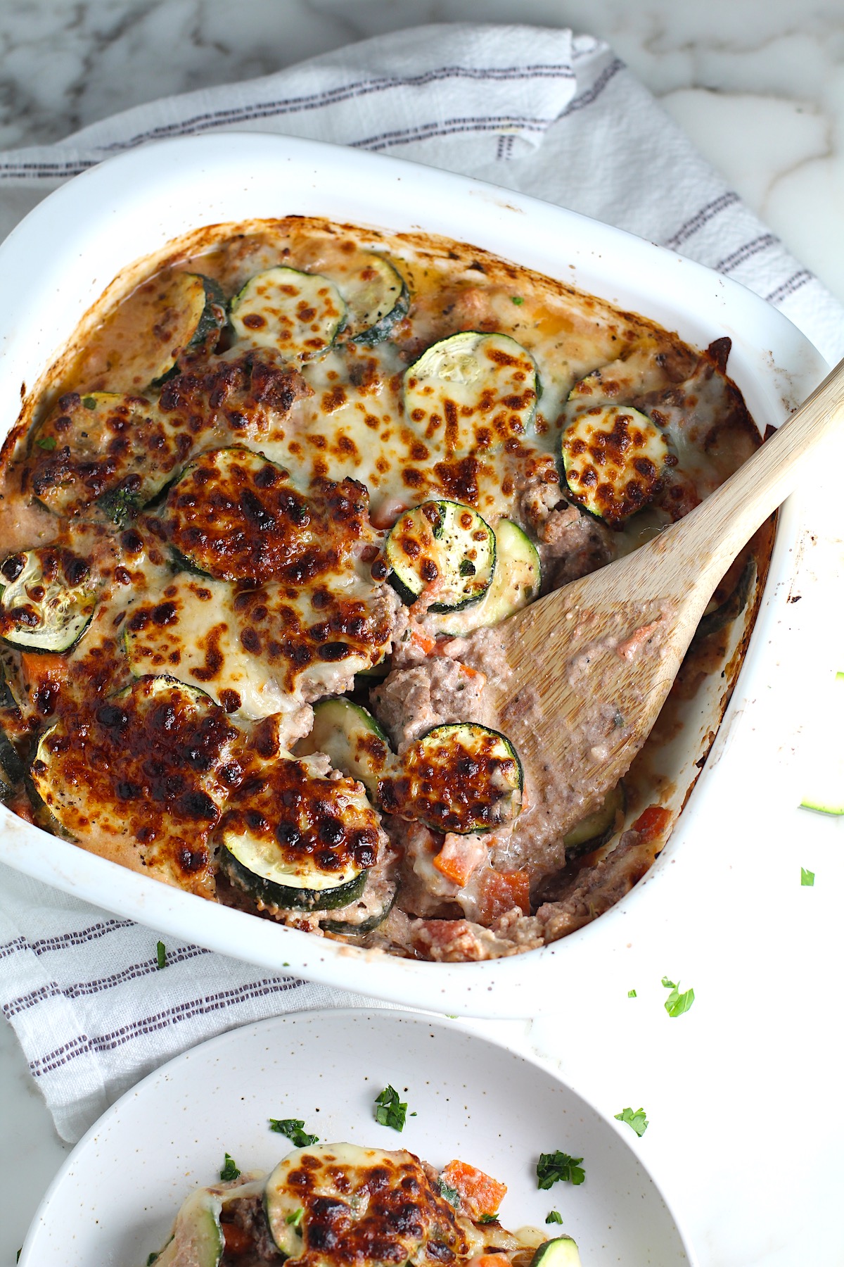 Ground Beef and Zucchini Casserole baked golden brown on top with wood spatula in corner of dish after some is scooped out. Sitting on blue and white stripped dish towel on counter with plate next to it.