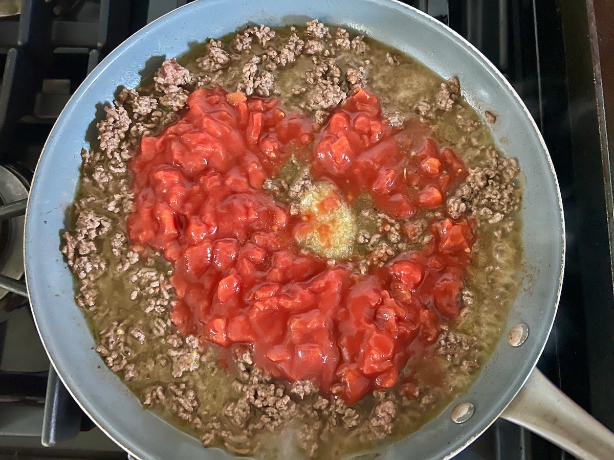 Ground beef cooking in a pan on the stove with diced tomatoes and minced garlic added for Ground Beef Gnocchi skillet recipe.