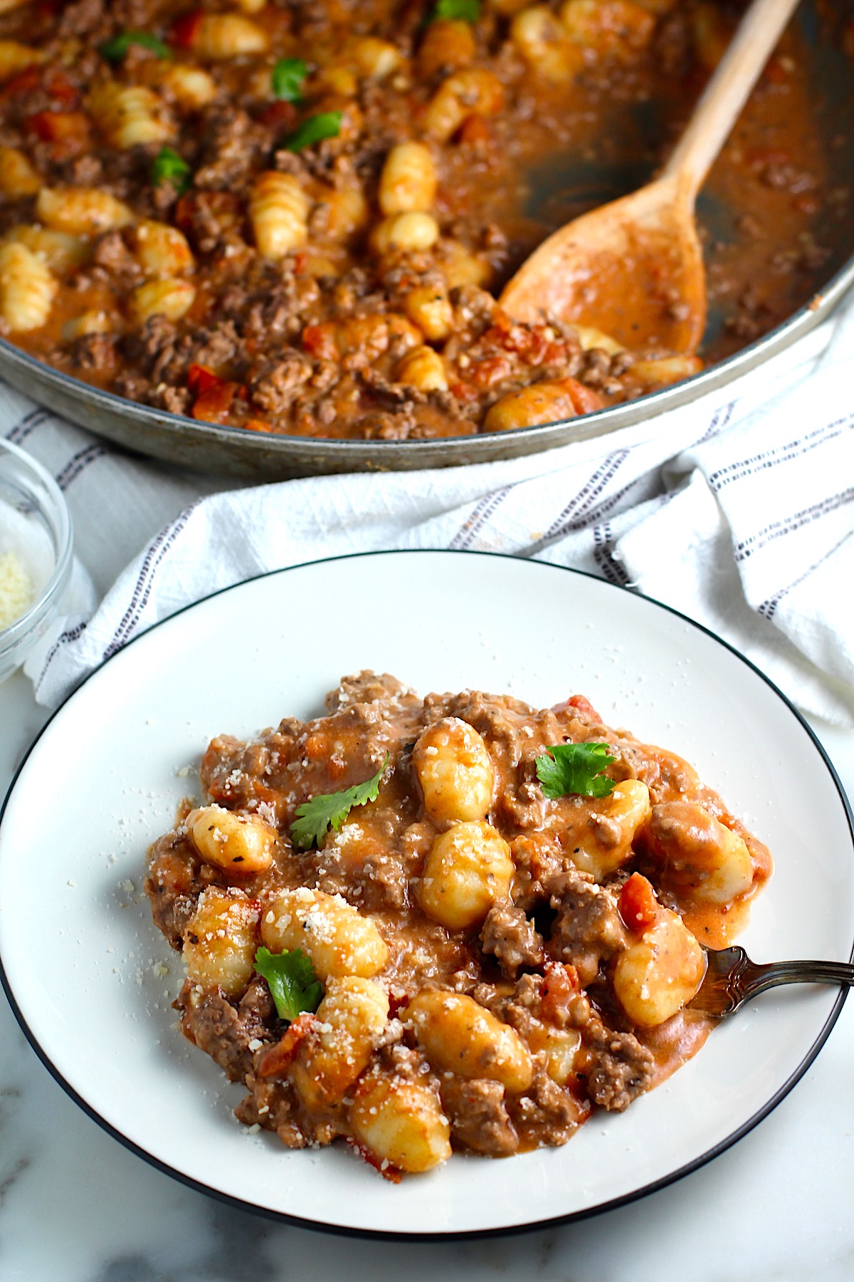Ground Beef Gnocchi Skillet recipe on a plate with fresh parsley garnish and a fork.  In the background is the skillet with the Beef Gnocchi and a wood spatula.