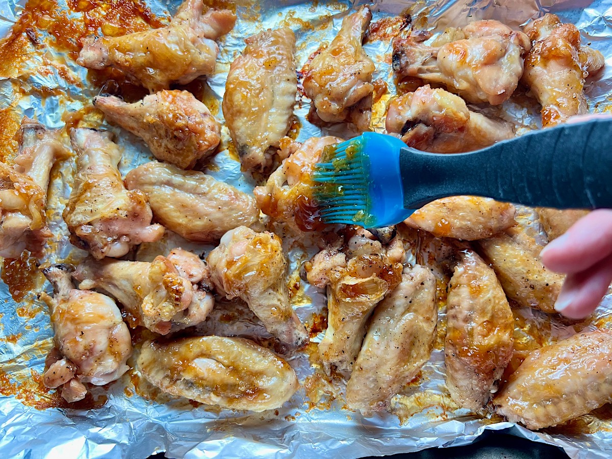 Blue silicone brush spreading Apricot sauce over cooked chicken wings and drumettes on a foil-lined sheet pan for the Apricot Sauce Wings recipe.