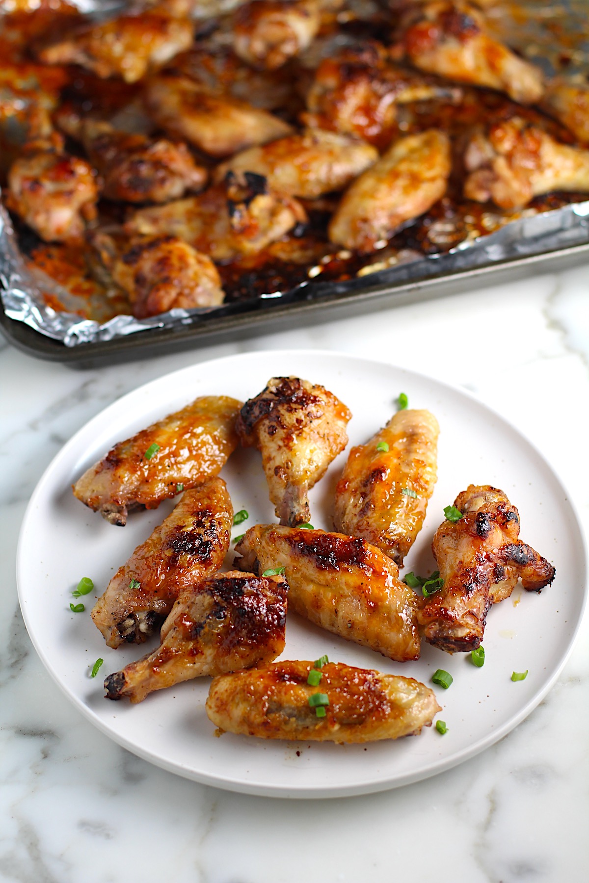 Sticky Garlic Soy Apricot Sauce Wings on a plate with scallion slices sprinkled on top and pan with more wings in background.