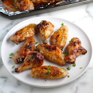 Sticky Garlic Soy Apricot Sauce Wings on a plate with scallion slices sprinkled on top and pan with more wings in background.
