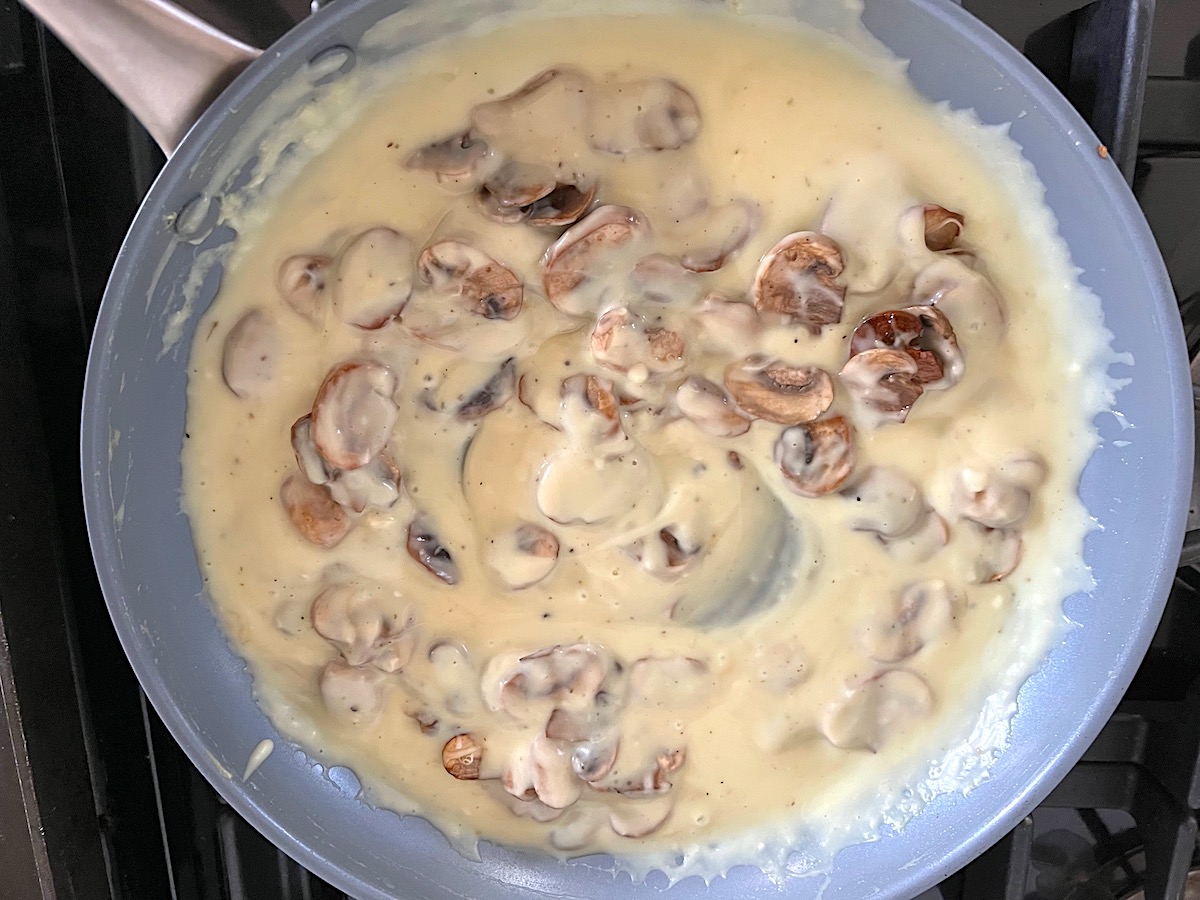 Mushrooms mixed into the alfredo sauce in a larger frying pan on the stove for Mushroom Alfredo Fettuccine pasta.