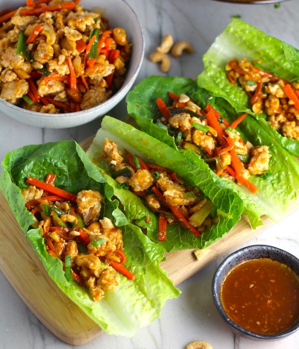 4 Cashew Chicken Lettuce Wraps on counter with a bowl of Cashew Chicken and Carrot filling on left and sauce on right.