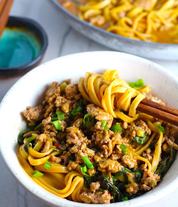 Dan Dan Noodle Recipe with Ground Chicken in a bowl. Chopsticks have noodles wrapped around and scallion slices are on top. The pan of noodles is in background.