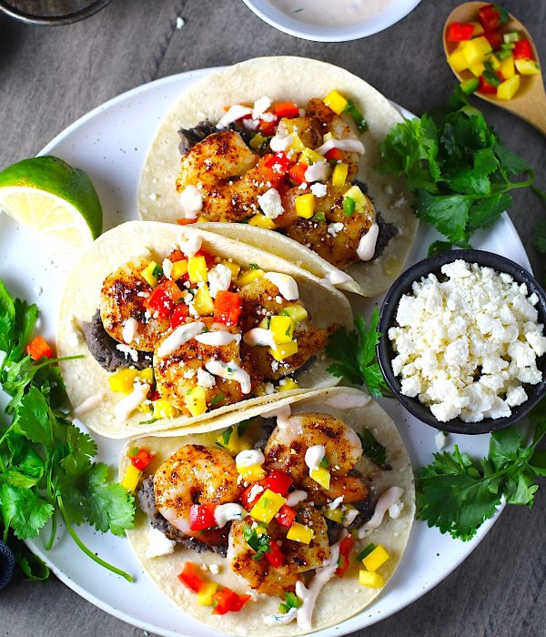 Grilled Shrimp Tacos in corn tortillas on a plate with Mango Salsa & Chipotle Crema. On the side are bowls of cotija cheese, crema, salsa, and cilantro leaves.